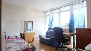 A bedroom with a bed, a desk, a chair, a dresser, a radiator, and a window with curtains. - 2-bed Apartment in London Woolwich (London)