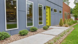 Een modern gebouw met grote ramen, een gele deur en een verzorgde tuin met vorm gesnoeide struiken en een grindpad.
