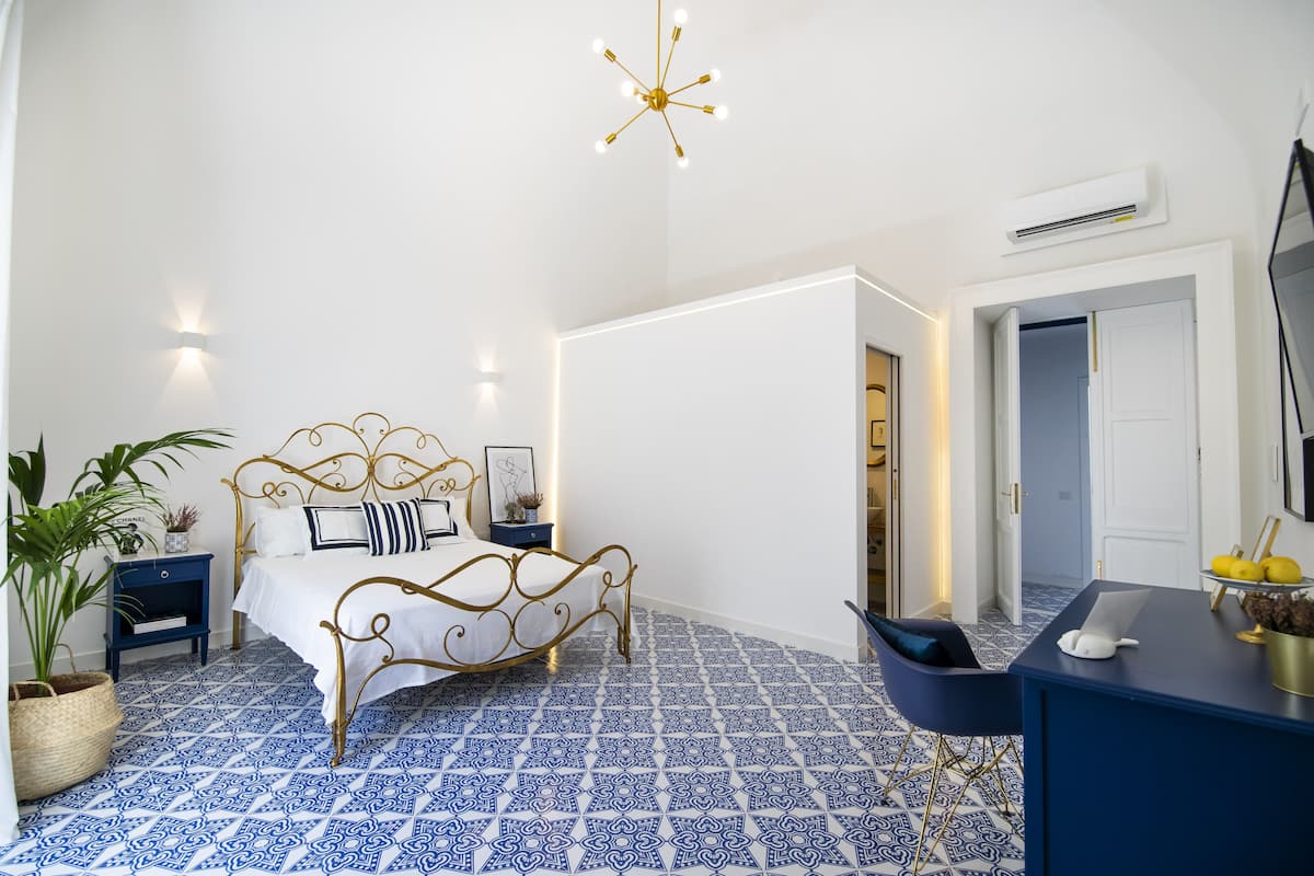 A bedroom with a bed, bedside tables, a desk, a chair, a potted plant, and a ceiling light fixture.