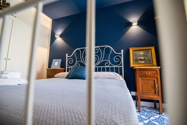 A neatly made bed with a white comforter and pillows, a wooden nightstand with a framed picture, and a wall-mounted light fixture. - Crudo Home (Pompei)