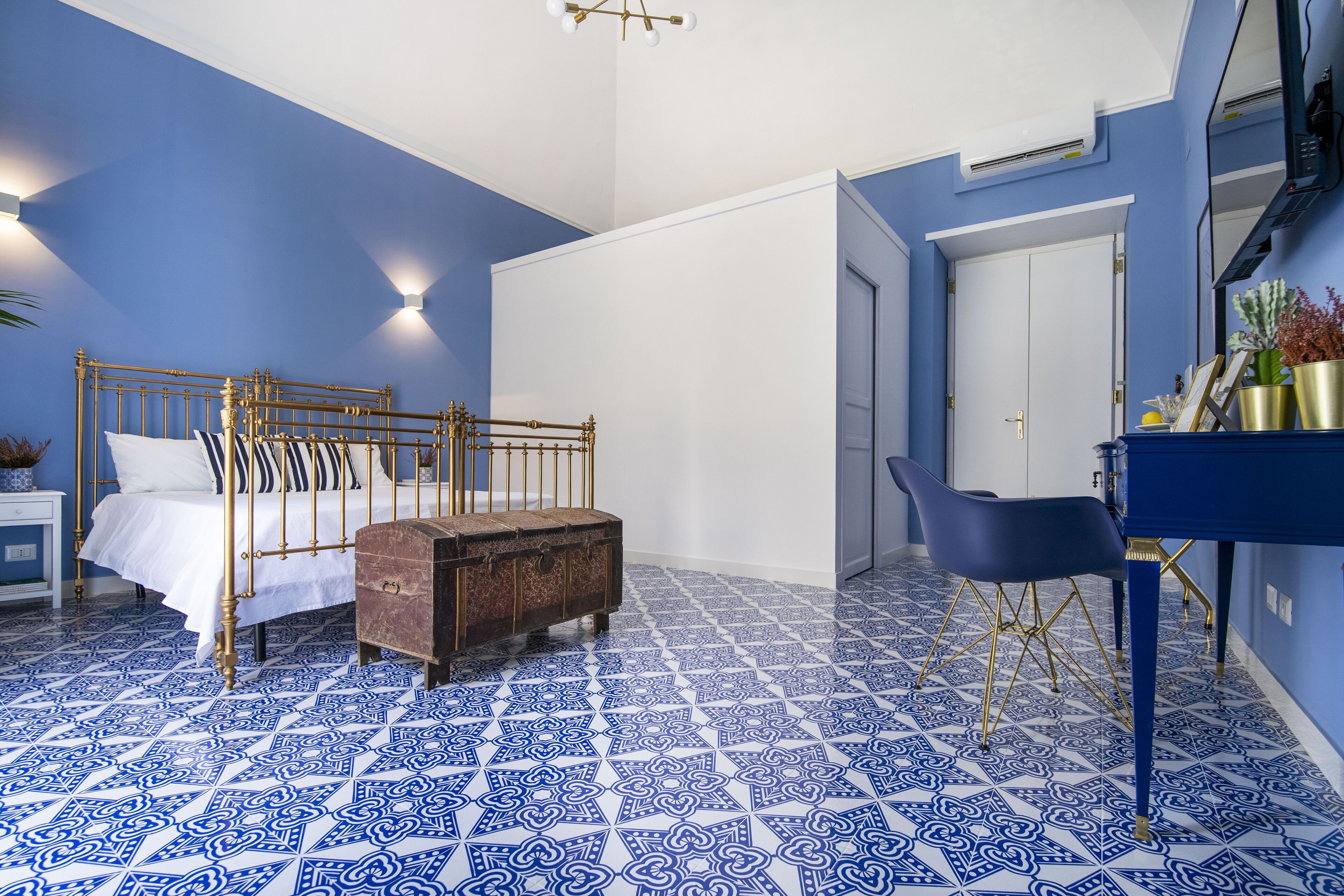 Un dormitorio con piso azul y blanco estampado, una cama con estructura dorada, un cofre de madera y un escritorio con una silla.
