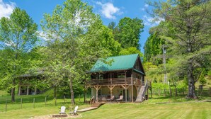 A wooden cabin with a green roof, surrounded by lush greenery and trees. - Woodsy Retreat with Hot Tub | Dogs Welcome | On River Time (Lansing)
