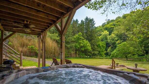A hot tub under a wooden pergola with a view of a lush green park. - Woodsy Retreat with Hot Tub | Dogs Welcome | On River Time (Lansing)