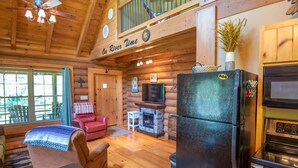 A log cabin interior with a dining area, a fireplace, a television, and a kitchen with a refrigerator and microwave. - Woodsy Retreat with Hot Tub | Dogs Welcome | On River Time (Lansing)