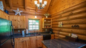 A log cabin kitchen with wooden cabinets, a granite countertop, a refrigerator, a sink, a window, and a chandelier. - Woodsy Retreat with Hot Tub | Dogs Welcome | On River Time (Lansing)