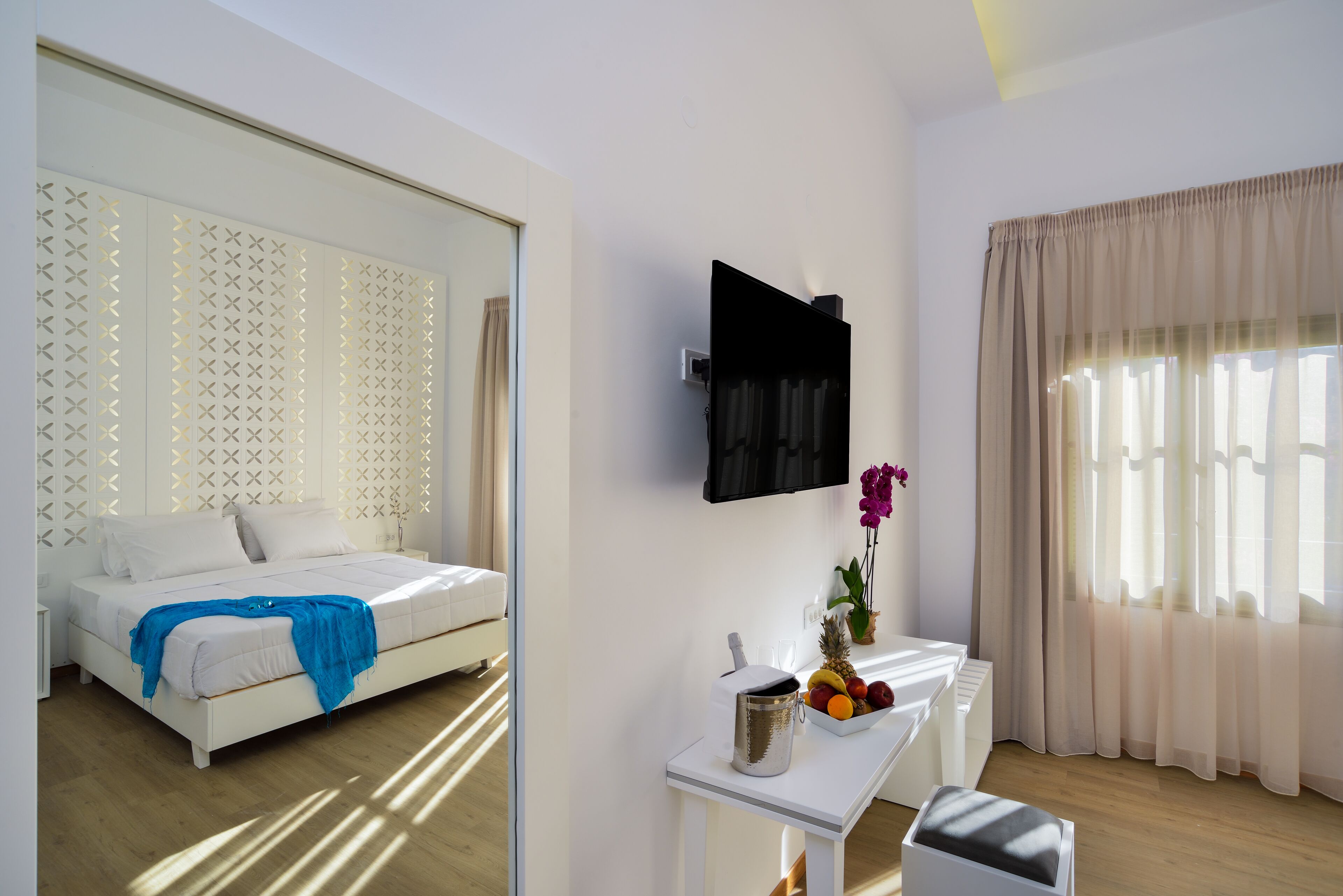 Ein modernes Hotelzimmer mit einem Bett, einem an der Wand montierten Fernseher, einem kleinen Tisch mit Obstschale und einem Fenster mit durchsichtigen Vorhängen.