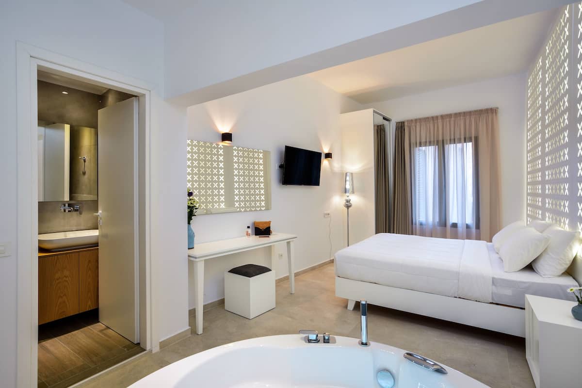 Een moderne hotelkamer met een groot, wit bad, een aparte badkamer met wastafel, een bureau met stoel, een bed met wit beddengoed en een aan de muur bevestigde televisie.