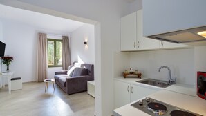 A modern kitchen with white cabinets, a built-in stove, and a sink. There is a gray sofa and a small white table in the adjacent living area. - Elia Portou Luxury Residence (Chania)