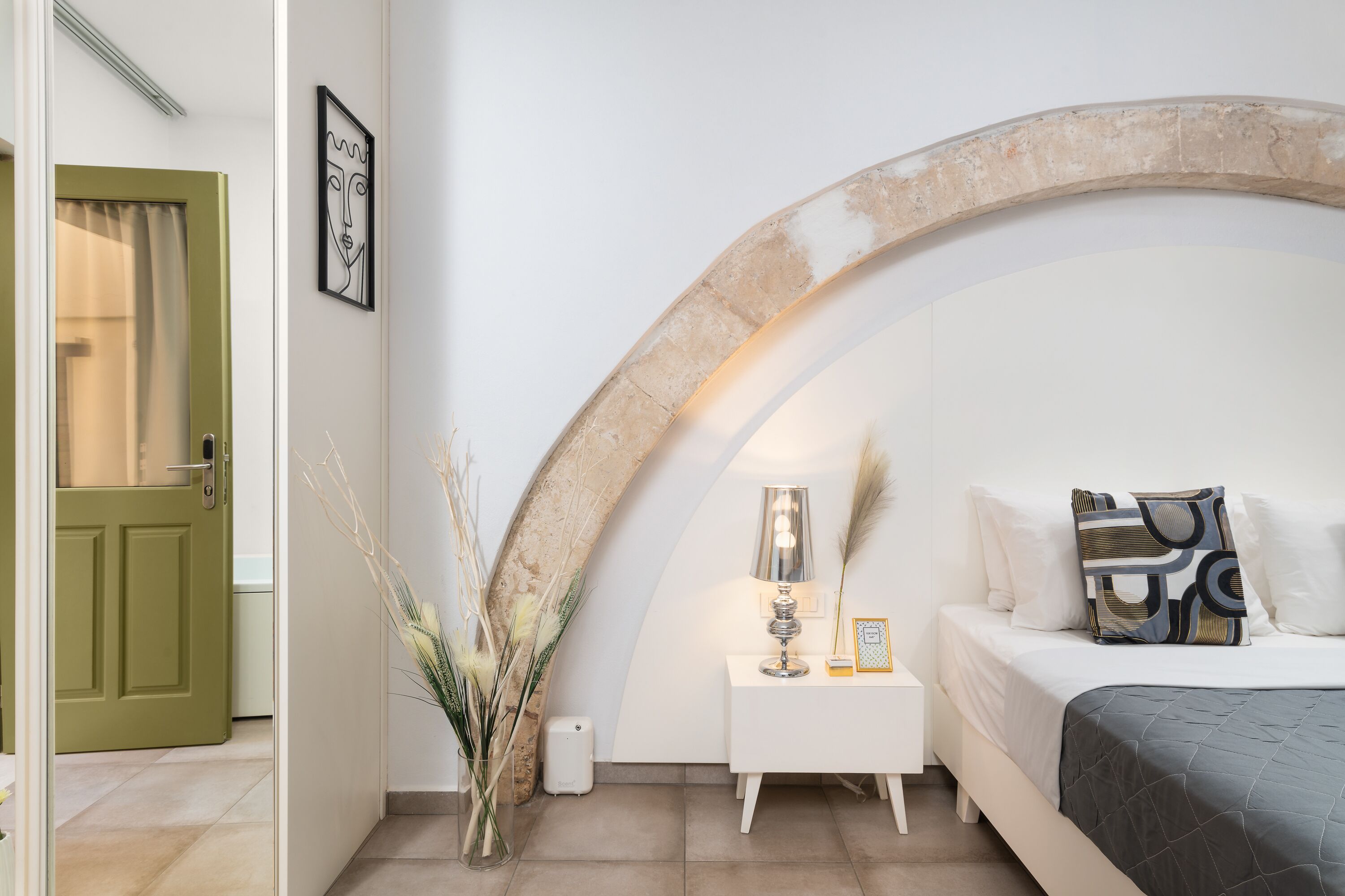 A bedroom with a bed, a bedside table with a lamp, a vase with dried plants, and a door with a green frame.
