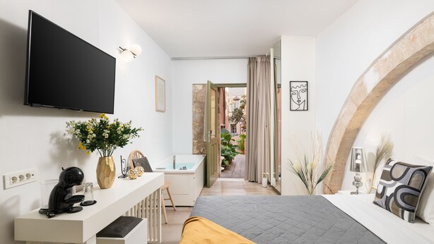 A modern bedroom with a flat-screen TV, a desk with a coffee machine, a bed with a quilted cover, and a bathroom visible through an open door. - Elia Portou Luxury Residence (Chania)