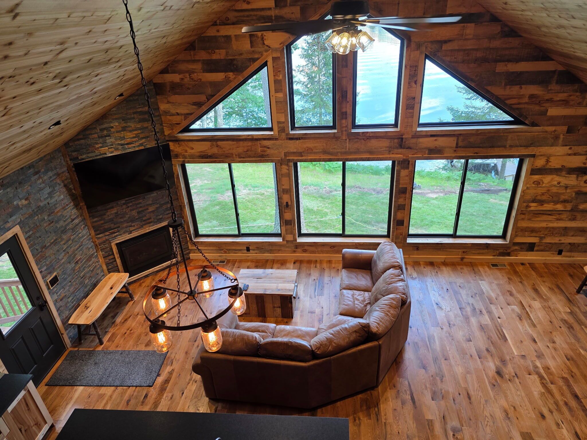 A cozy cabin interior with a loft-style ceiling, a sectional sofa, a fireplace, and large windows offering a view of the outdoors.