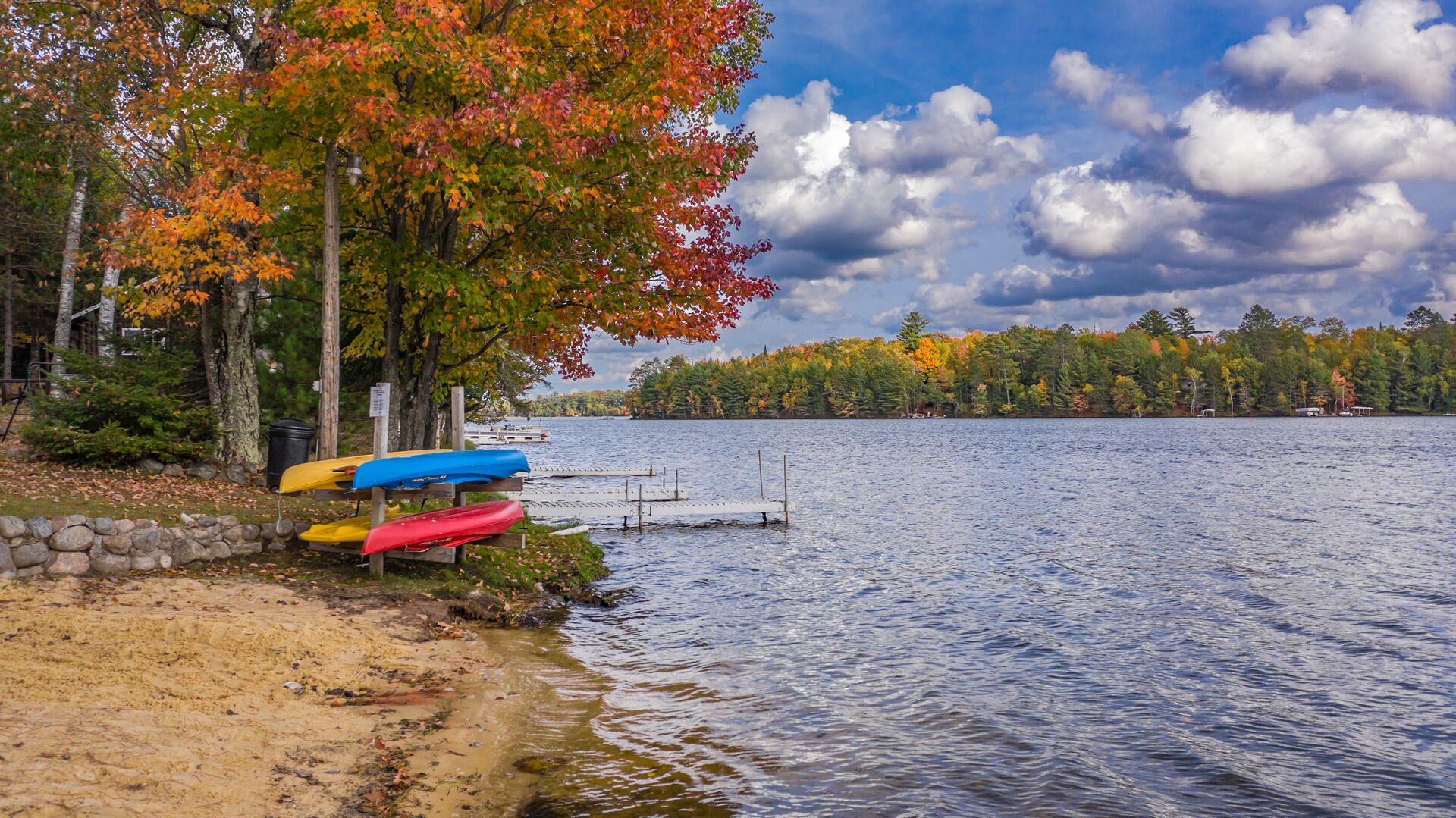 Escena junto al lago con kayaks, un muelle y follaje otoñal.