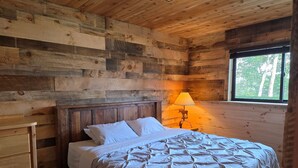 A wooden cabin bedroom with a bed, a nightstand, a dresser, and a window with a view of trees. - Brand new pet friendly vacation home with private dock and firepit! (St. Germain)