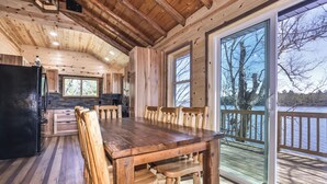 A wooden cabin interior with a dining table and chairs, a kitchen area, and a large window overlooking a body of water. - Remodeled lakeside resort cabin with fireplace, sandy beach, dock, firepit! (St. Germain)