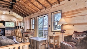 A cozy cabin interior with wooden walls and a high ceiling. There is a dining table with chairs, a sofa, and a lamp. Large windows provide views of the outdoors. - Remodeled lakeside resort cabin with fireplace, sandy beach, dock, firepit! (St. Germain)