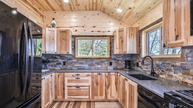 A kitchen with wooden cabinets, a stone backsplash, a built-in oven, and a window overlooking greenery. - Remodeled lakeside resort cabin with fireplace, sandy beach, dock, firepit! (St. Germain)