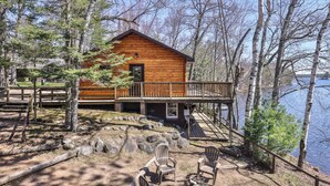 A wooden cabin with a deck and a view of a lake. - Remodeled lakeside resort cabin with fireplace, sandy beach, dock, firepit! (St. Germain)
