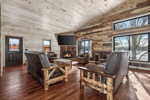 A log cabin interior with a fireplace, wooden furniture, and large windows offering a view of the outdoors.