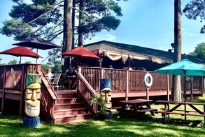 Una terraza de madera con mesas de picnic y sombrillas, rodeada de árboles.
