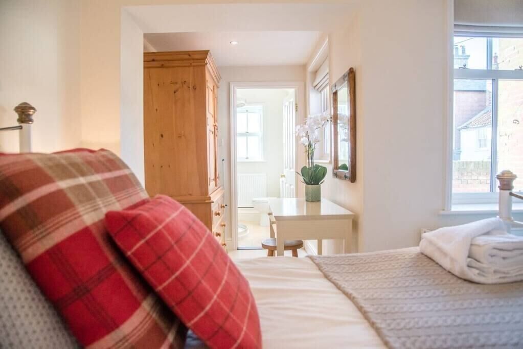 A bedroom with a bed, a wooden wardrobe, a desk with a chair, and a window with a view of the outside.