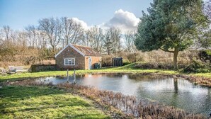 A rustic brick house with a pond in front, surrounded by trees and a grassy area. - Mill Cottage, Tunstall (Suffolk)