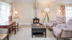 A cozy living room with a fireplace, wooden furniture, and floral curtains. - Mill Cottage, Tunstall (Suffolk)