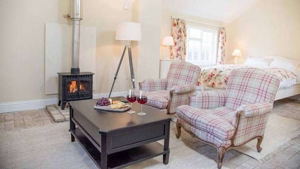 A cozy room with a fireplace, plaid chairs, a table with glasses and grapes, and a bed with floral curtains. - Mill Cottage, Tunstall (Suffolk)