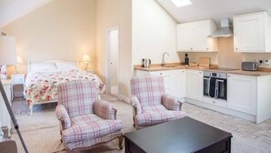 A compact bedroom with a bed, two chairs, and a small table. Adjacent is a kitchenette with a stove, oven, and sink. - Mill Cottage, Tunstall (Suffolk)