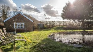 A rustic stone house with a sloped roof, a small pond, and a grassy area with lounge chairs. - Mill Cottage, Tunstall (Suffolk)