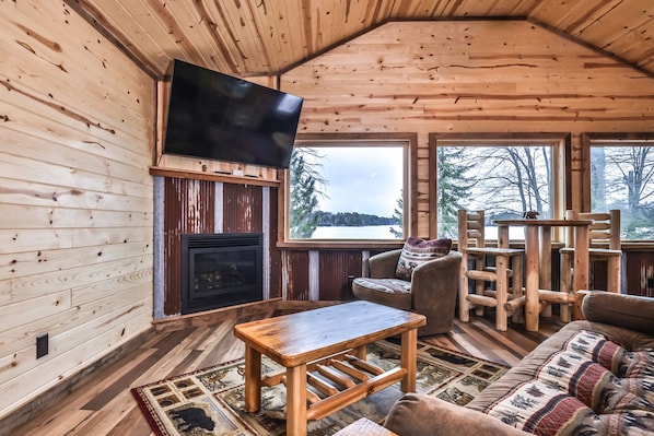 L’intérieur chaleureux d’un chalet, avec une cheminée, des meubles en bois et une grande fenêtre donnant sur des arbres et un lac.