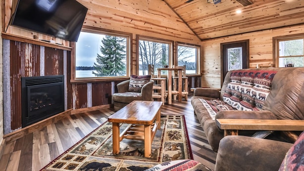 Interior acogedor de una cabaña con chimenea, muebles de madera y grandes ventanales que ofrecen vistas al exterior.