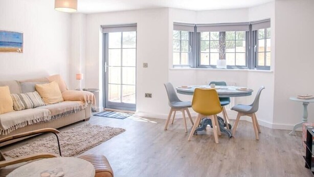 A modern living room with a sofa, a dining table with chairs, and a large window with a view. - 3 Oaks Court, Thorpeness (Leiston)