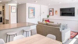 A modern kitchen with a central island, a dining area with a table and chairs, a sofa with cushions, and a flat-screen TV mounted on the wall.