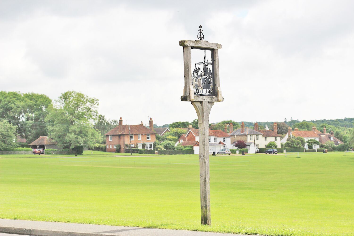 A wooden signpost with a coat of arms and the name "WOODCHURCH" in the center, set against a backdrop of a green field and houses.