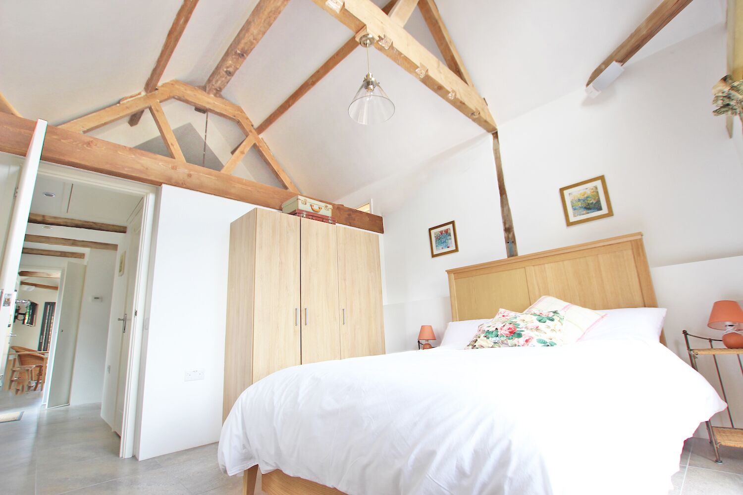 A bedroom with a wooden ceiling, a bed with white bedding, a wooden wardrobe, and a small table with a lamp.