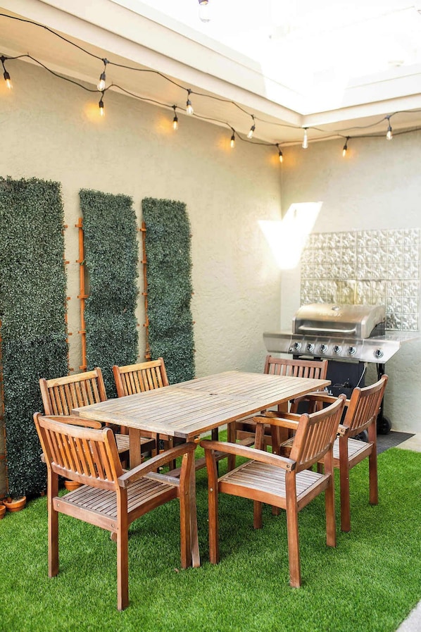 Um pátio externo com mesa e cadeiras de madeira, churrasqueira e luzes decorativas em fio.