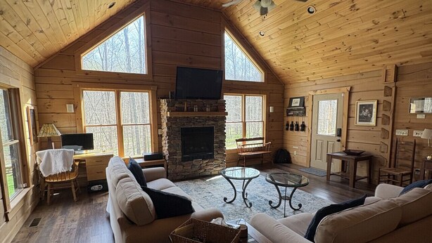 A log cabin interior with a stone fireplace, two sofas, a dining table, and a ceiling fan.