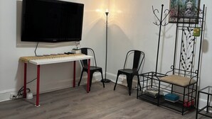 A room with a flat-screen TV, a white desk, two chairs, a floor lamp, and a decorative wall piece.