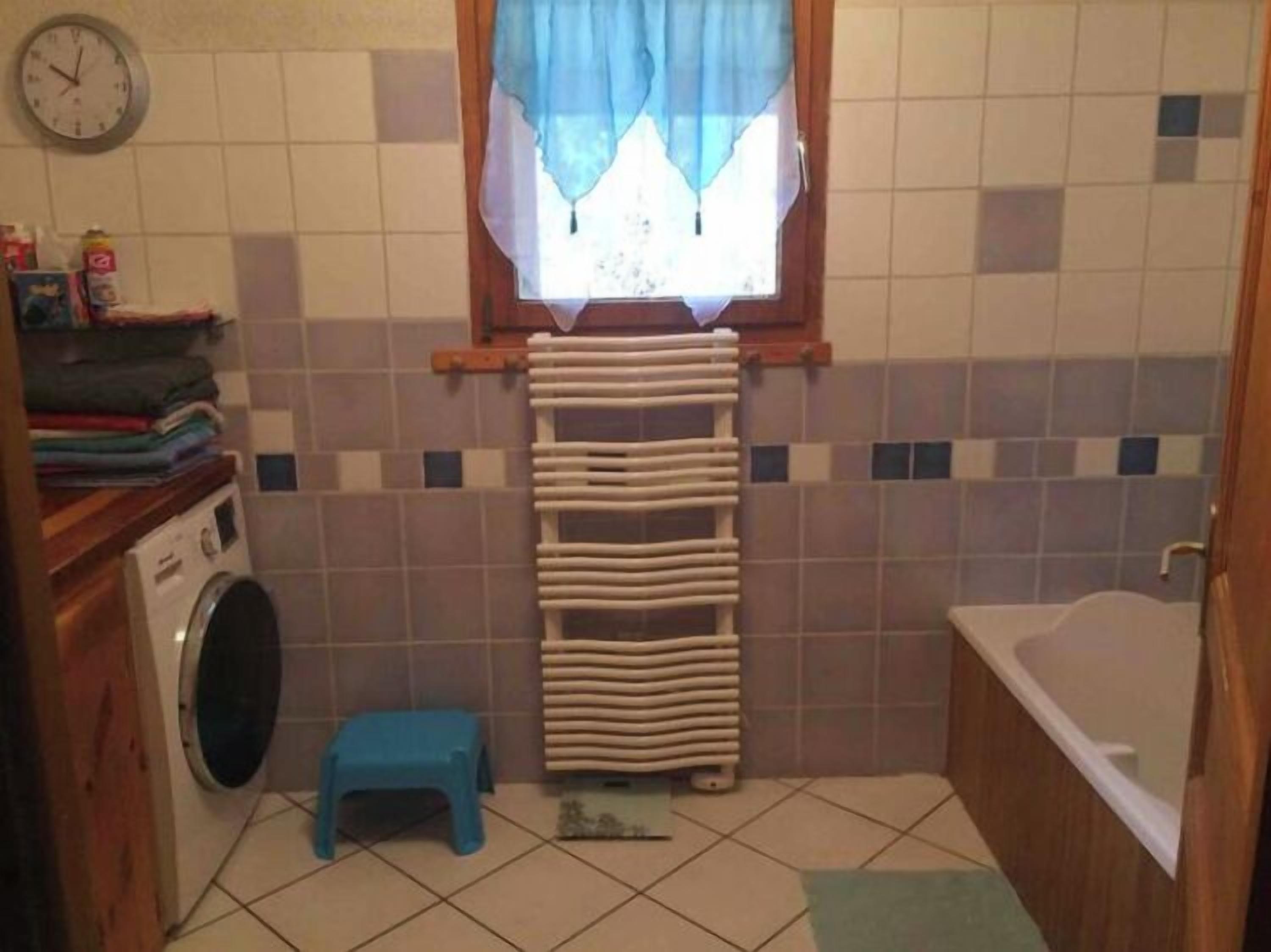 A small bathroom with a washing machine, a blue stool, a towel rack with folded towels, a clock on the wall, and a bathtub.