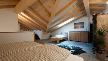 Ein Schlafzimmer mit schrägen Wänden, einem Bett mit karierter Bettdecke, einem kleinen Nachttisch, einem Beistelltisch, einem Holzschrank und einer Zimmerpflanze.