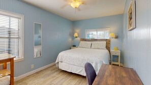 A bedroom with a bed, bedside lamps, a wooden headboard, a nightstand, a chair, a wooden table, a ceiling fan, and a window with blinds.