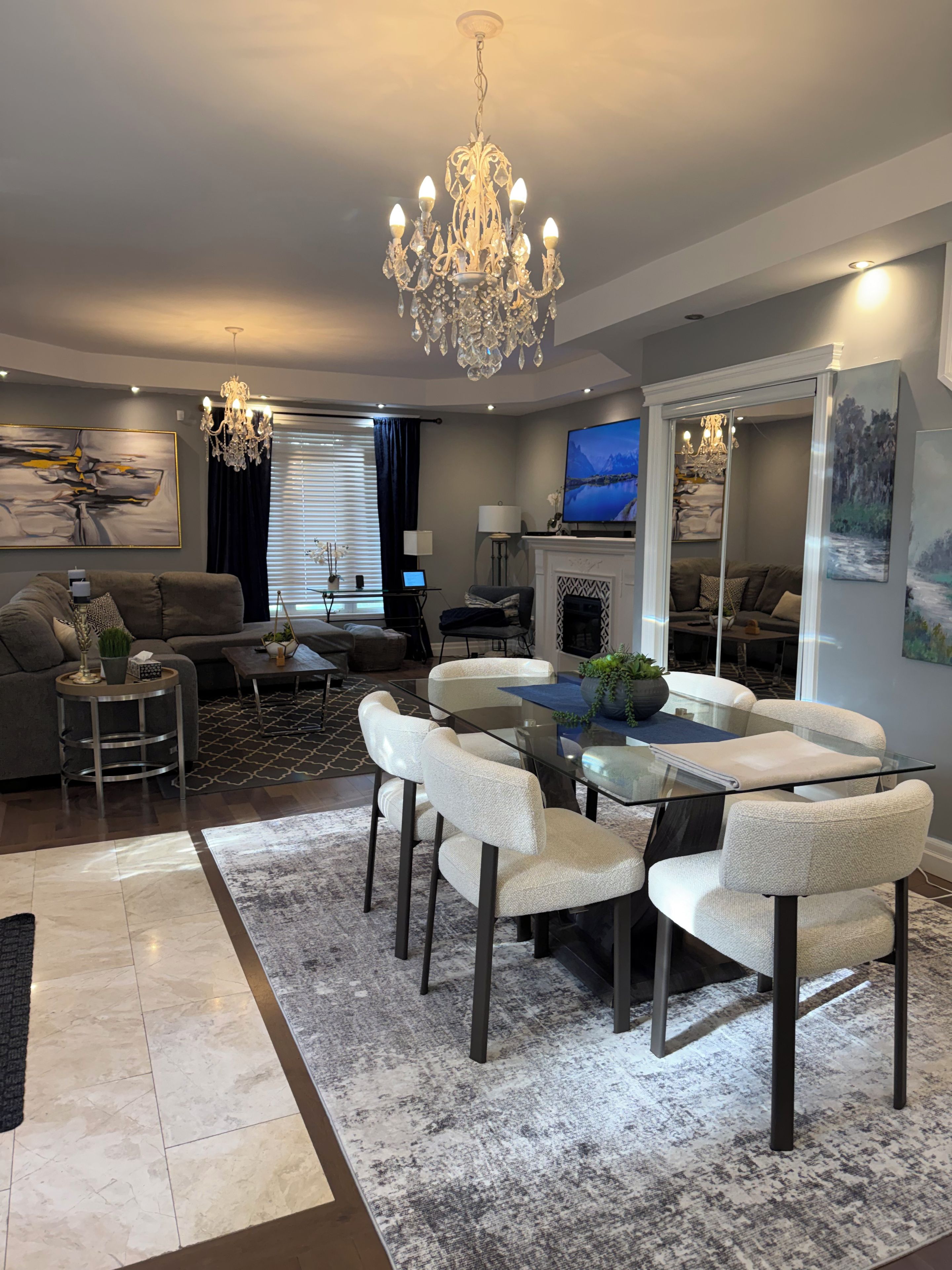 A modern living room with a glass dining table, white upholstered chairs, a chandelier, a fireplace, and a large painting on the wall.