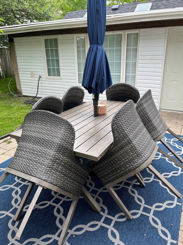 A patio with a wooden table and wicker chairs, a blue umbrella, and a patterned rug.