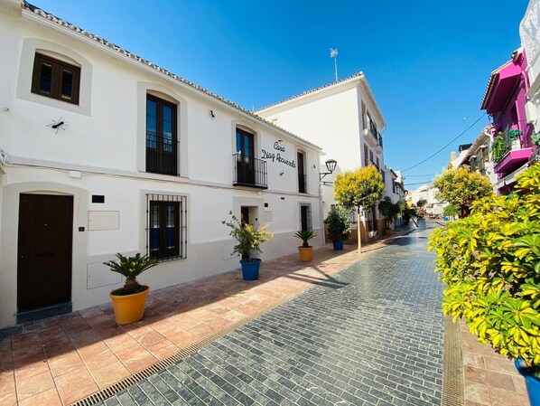 A narrow street with white buildings, a sign reading "Casa Di Pig Recuerda," and potted plants along the sidewalk.