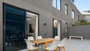 A modern patio with a wooden table and white chairs, large glass sliding doors, and a grey exterior wall. - Chic Green Point Apartment | Fast WiFi | BBQ Patio (Cape Town)