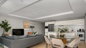 A modern living room with a grey sofa, a glass dining table, and a kitchen area with white cabinets and appliances. - Stunning Green Point Apartment , Patio + Fast WiFi (Cape Town)