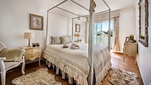 A four-poster bed with a canopy, a wooden nightstand with a lamp, a chair, and a large window with curtains. - Rosal 27 (Albufeira)