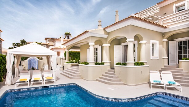 A swimming pool with lounge chairs, a covered seating area, and a building with arches and steps. - Rosal 27 (Albufeira)