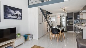 Uma sala de estar moderna com uma TV de tela plana, um espaço de jantar com mesa e cadeiras, uma escada que leva a um andar superior e uma cozinha com eletrodomésticos de aço inoxidável.