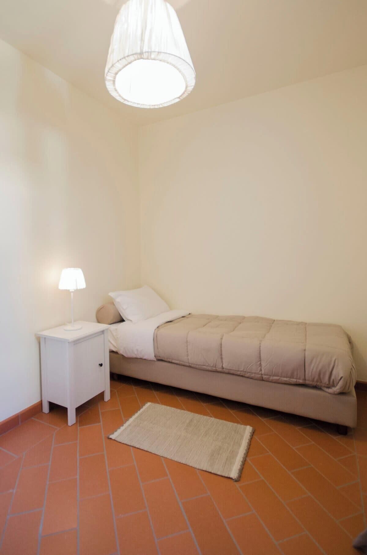 Una habitación pequeña con cama individual, una mesita de noche, una lámpara y una alfombra sobre suelo de baldosas.
